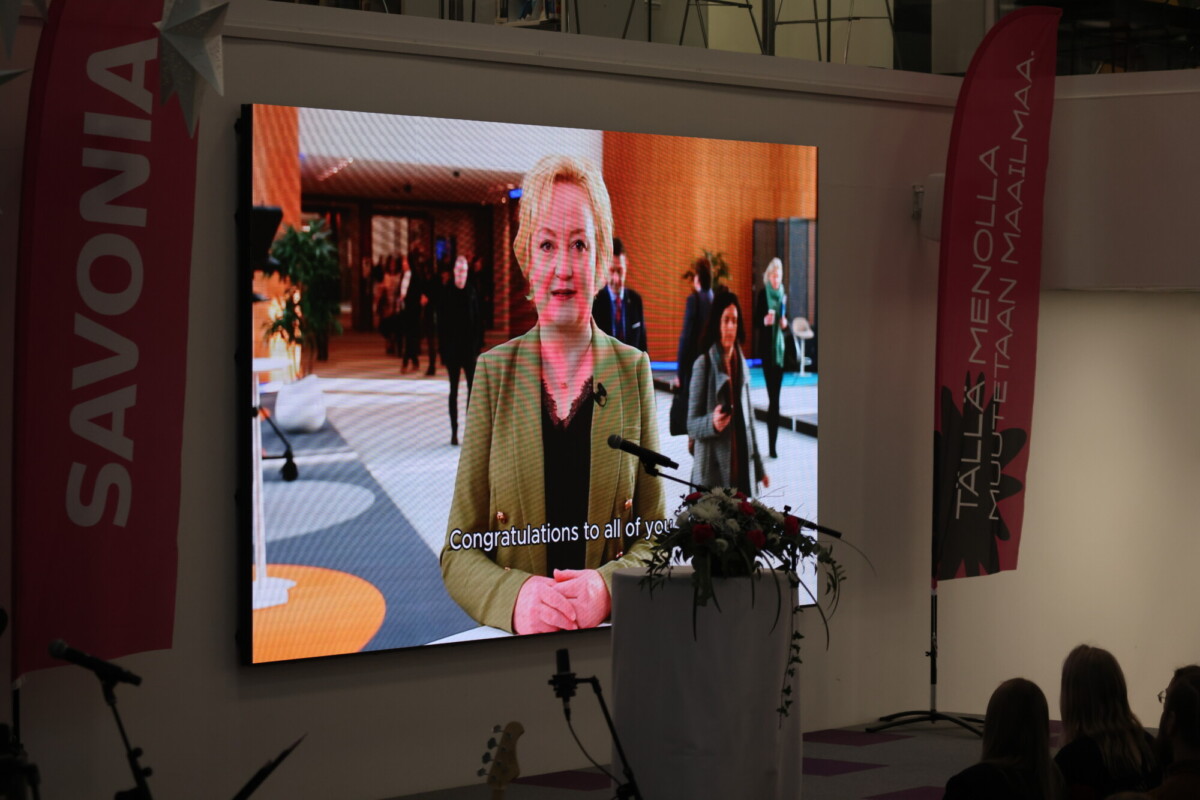 Sisätiloissa järjestettävässä tapahtumassa suurella näytöllä näkyy nainen puhumassa, ja tekstitys kuuluu: "Onnittelut teille kaikille." Näytön vieressä näkyy vaaleanpunaisia banderolleja, joissa on SAVONIA ja suomenkielinen teksti. Edessä on koroke, jossa on kukkia.