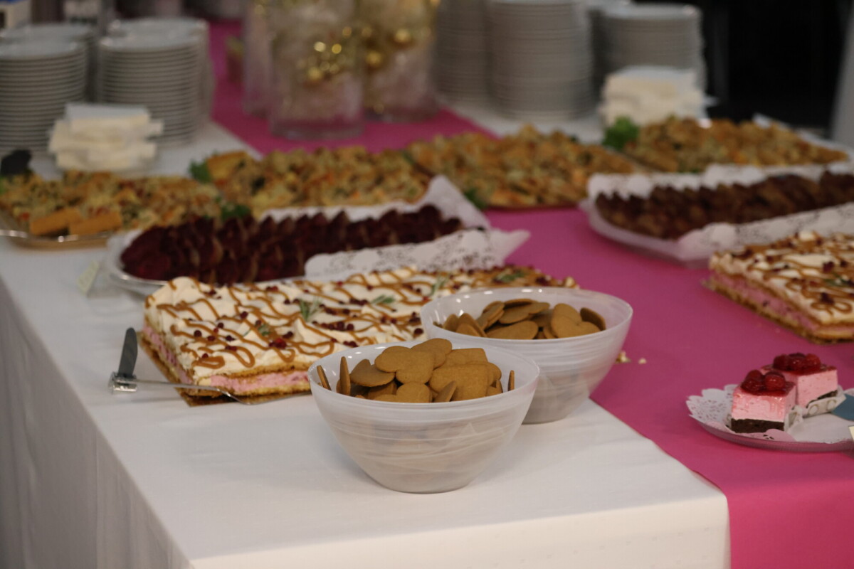 Jälkiruokapöytä, joka on peitetty vaaleanpunaisella ja valkoisella kankaalla, ja jossa on kulhoja keksejä, erilaisia kakkuja ja leivonnaisia, taustalla lautaset ja lautasliinat pinottuna.