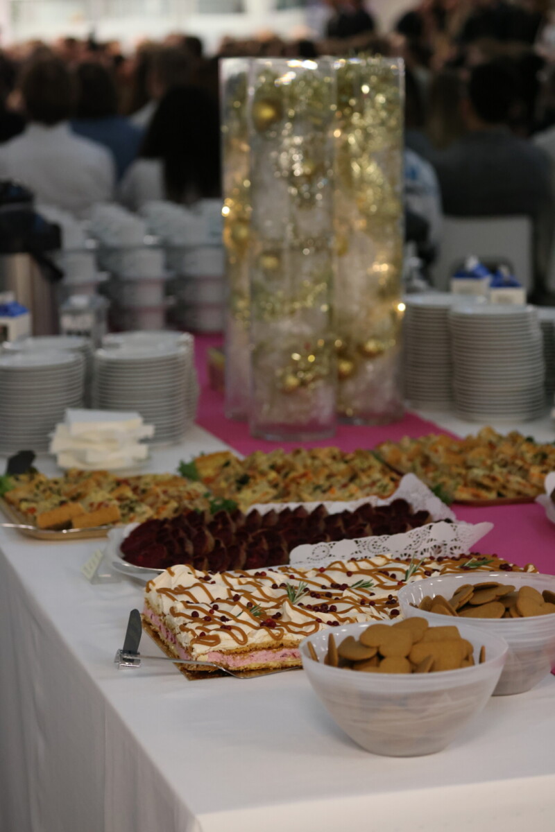 Pöytä, jossa on jälkiruokalautasia ja keksejä, joita ympäröivät pinotut valkoiset astiat ja kupit. Taustalla on korkea koristeellinen keskipiste, jossa on kultaisia koristeita, ja joukko ihmisiä istumassa.