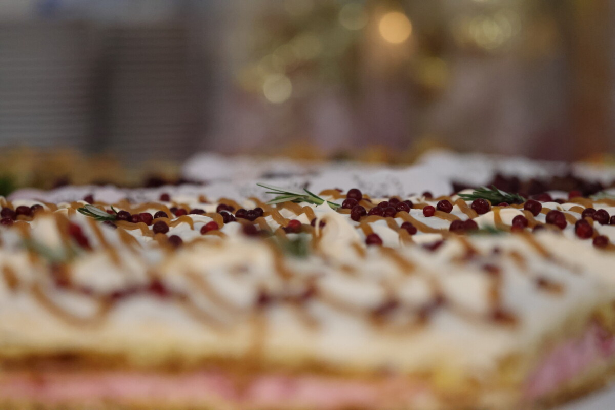 Lähikuva koristellusta kakusta, jossa on valkoinen kuorrutus, punaisia marjoja muistuttavia koristeita ja rosmariinin oksia, taustan ollessa sumea.