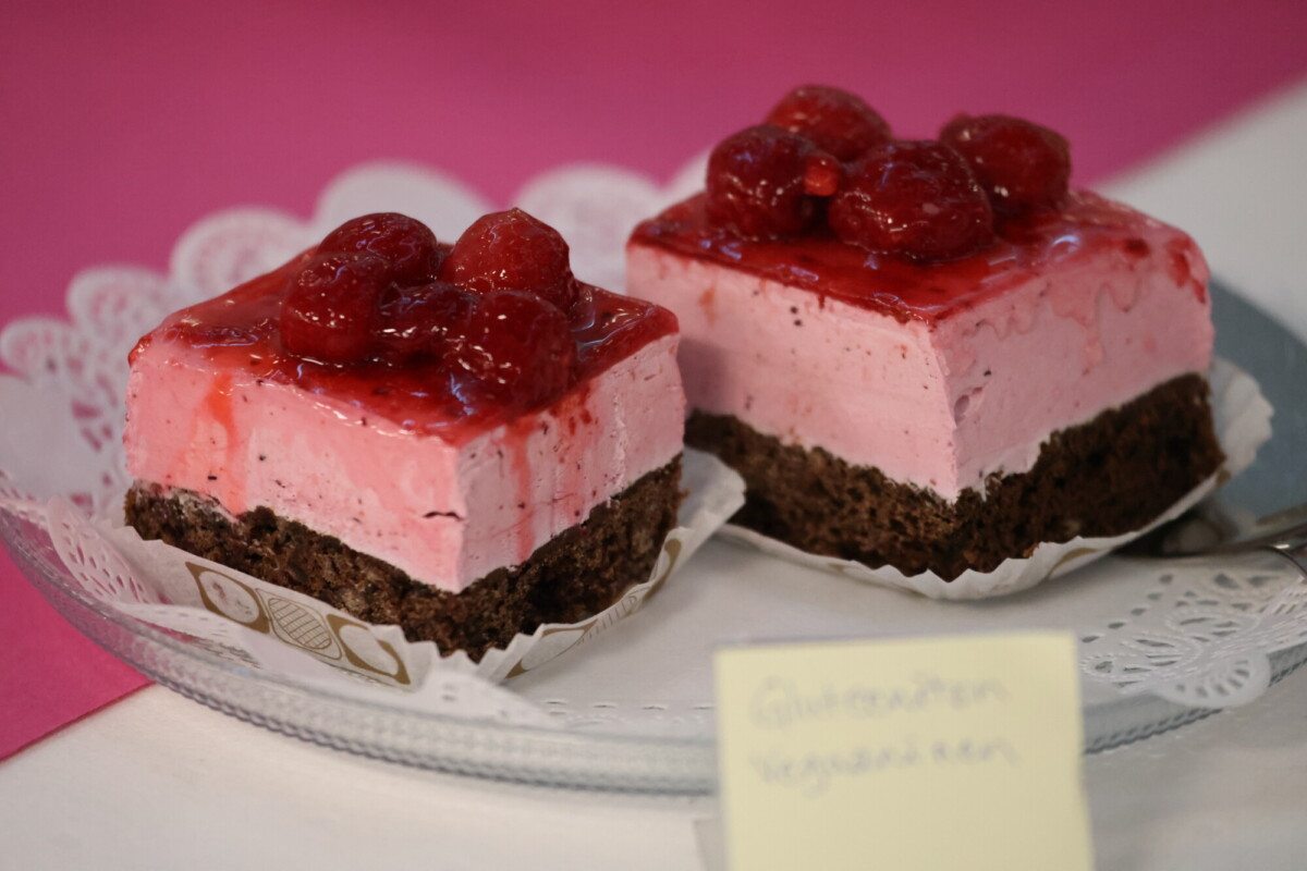 Kaksi palaa mansikkamousse-kakkua, jossa on suklaapohja ja jonka päällä on kerros kuorrutettuja mansikoita, on esillä lasilautasella, jossa on käärinliina, ja sen vieressä on käsin kirjoitettu keltainen viesti.