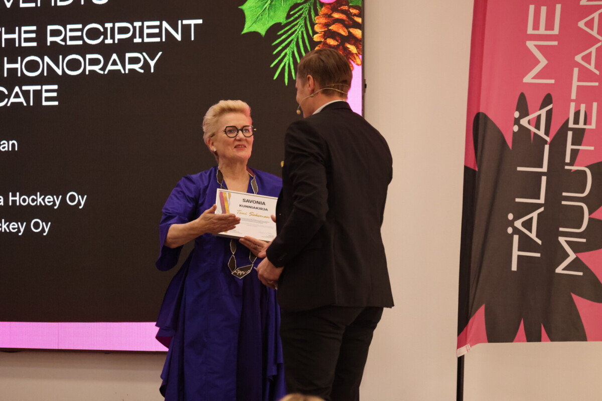 Siniseen mekkoon pukeutunut nainen saa lavalla kunniakirjan pukumieheltä. Taustalla näkyy valkokangas ja suomenkielistä tekstiä sisältävä banderolli.