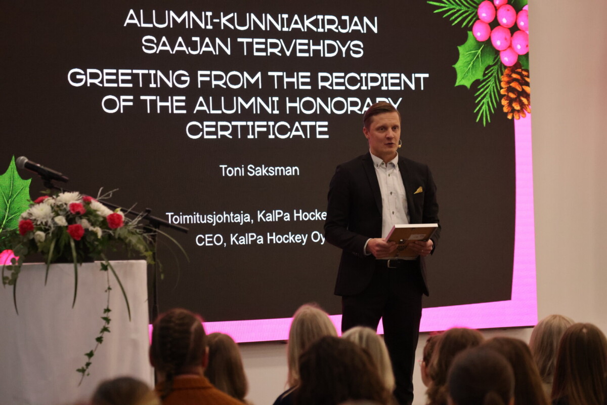 Pukumies pitää muistolaattaa ja puhuu lavalla näytön edessä, jossa näkyy loma-aiheinen viesti alumneille. Etualalla näkyy yleisö ja läheisellä korokkeella on kukka-asetelma.