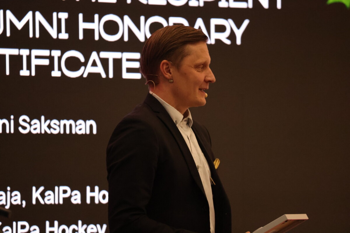 Pukumies seisoo lavalla mustaa taustaa vasten, jossa on suuri valkoinen teksti, suorakulmainen esine kädessään ja hymyilee, ilmeisesti esitelmän tai palkintoseremonian aikana.