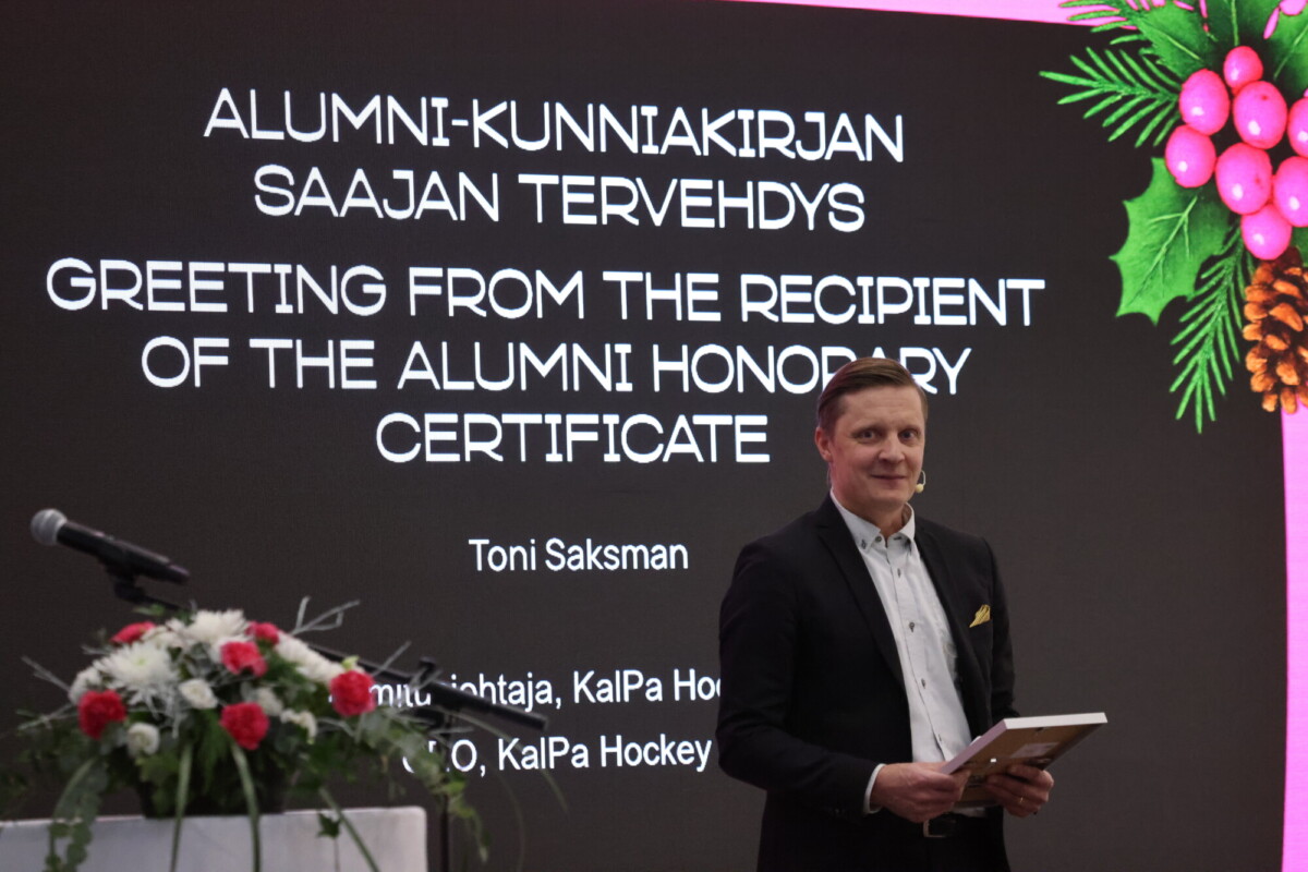 Pukumies seisoo lavalla mikrofonin ja kukka-asetelman vieressä muistolaatta kädessään. Hänen takanaan näytöllä näkyy kaksikielinen tervehdys ja hänen nimensä, Toni Saksman, juhlallisten koristeiden alla.