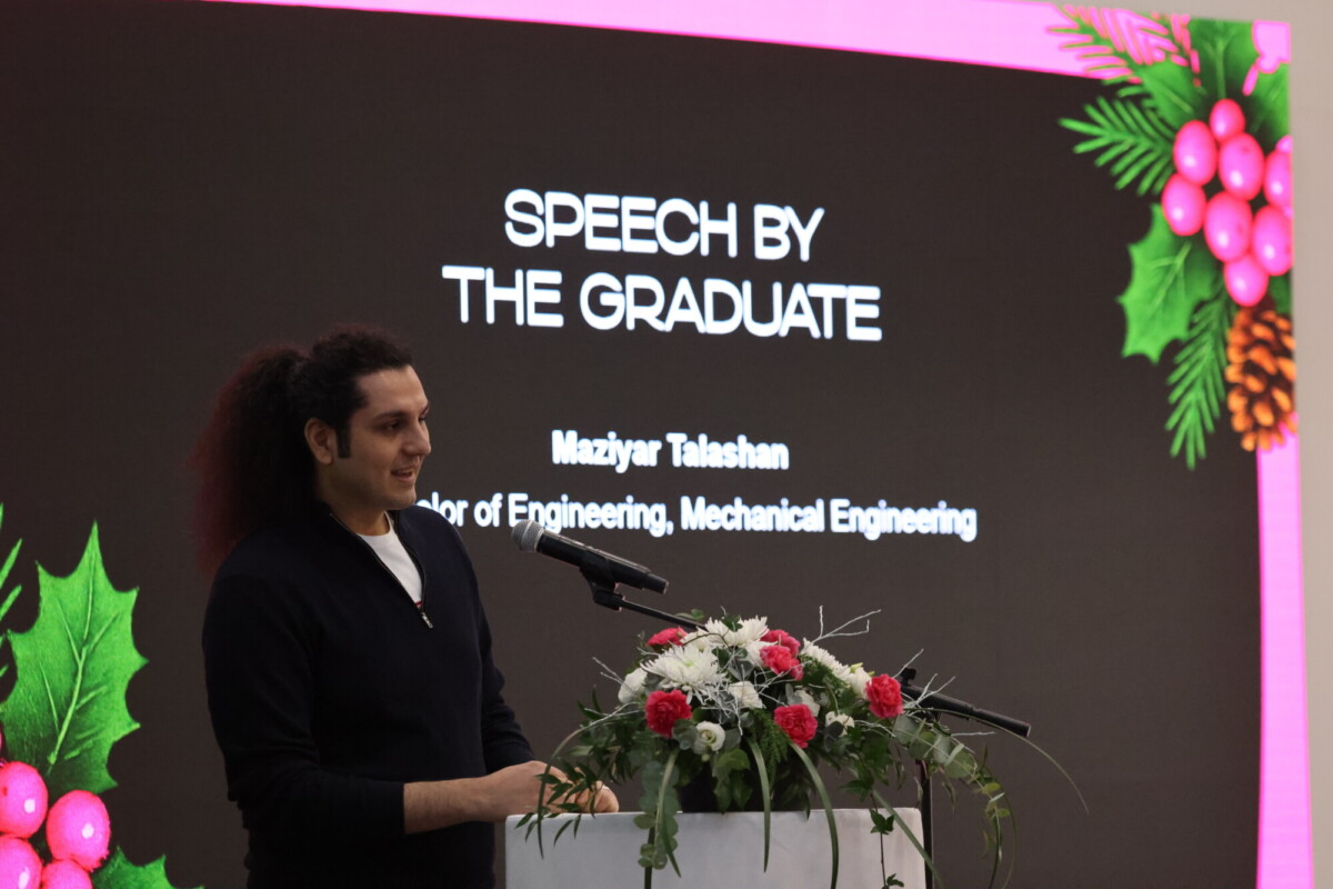 Mies seisoo kukkien koristelemalla korokkeella ja pitää puhetta. Hänen takanaan näytöllä lukee SPEECH BY THE GRADUATE, jossa on juhlallisia koristeita ja nimi, joka osoittaa, että hän on tekniikan kandidaatti.