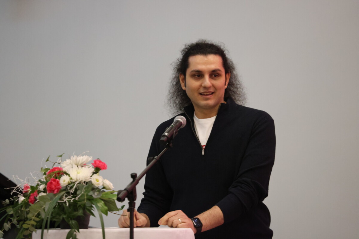 Pitkät tummat hiukset omaava henkilö puhuu korokkeella, jossa on mikrofoni, ja hänellä on musta villapaita ja kello. Korokkeella on kukka-asetelma, jossa on valkoisia ja vaaleanpunaisia kukkia. Tausta on yksivärinen ja vaalea.