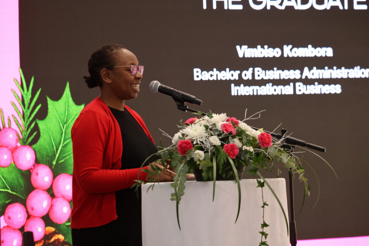 Punatakkinen nainen puhuu valkoisilla ja vaaleanpunaisilla kukilla koristellulla korokkeella. Hänen takanaan olevalla diaesityksellä näkyy The Graduate, hänen nimensä ja akateeminen ohjelma.