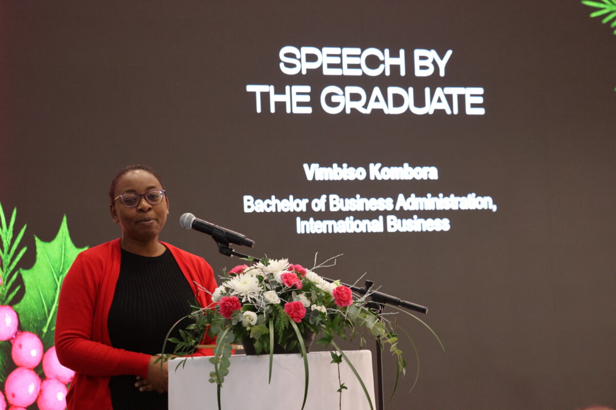 Punatakkinen nainen seisoo kukkien koristelemalla korokkeella ja puhuu mikrofoniin. Hänen takanaan näytöllä lukee Speech by the Graduate, Vimbiso Kombora, Bachelor of Business Administration, International Business.
