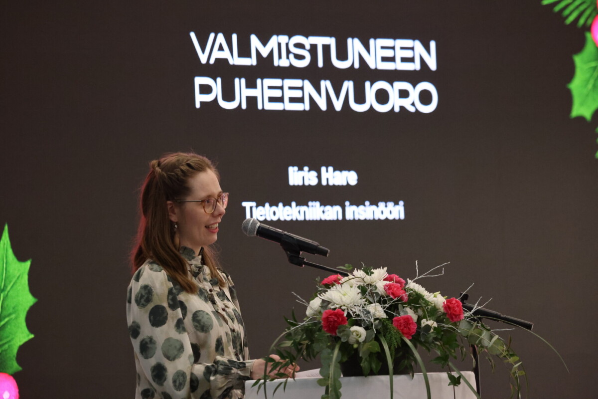 Nainen puhuu kukilla koristellulla korokkeella, edessä on näyttö, jossa näkyy teksti VALMISTUNEEN PUHEENVUORO, Iiris Häre, Tietotekniikan insinööri, näytöllä on vihreitä ja punaisia koristeita.