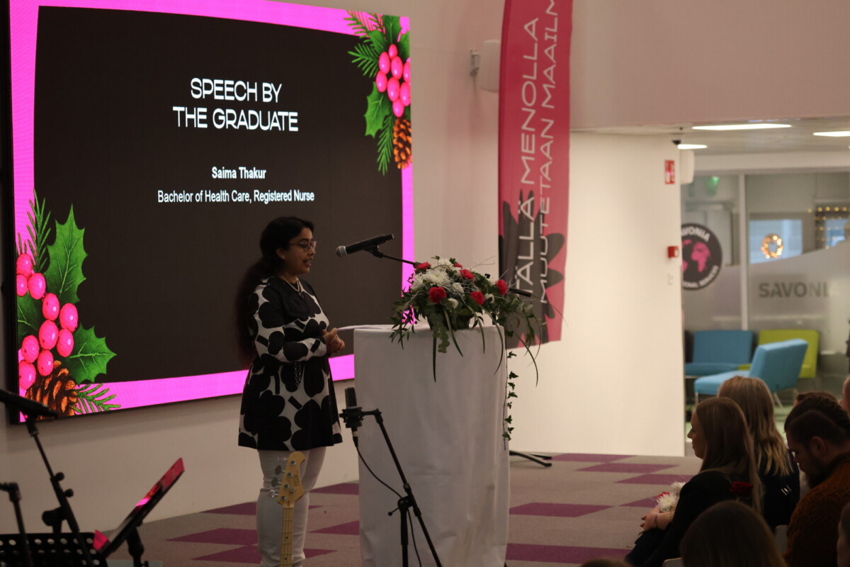 Nainen seisoo kukkien koristelemalla korokkeella ja pitää puhetta yleisölle. Hänen takanaan näytöllä lukee: SPEECH BY THE GRADUATE: Saima Thakur, Bachelor of Health Care, Registered Nurse, juhlallisin pyhimyskoristein.