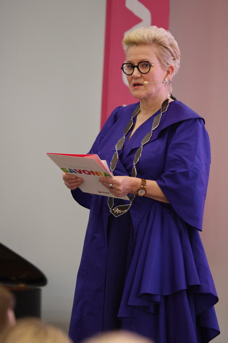 Lyhyet vaaleat hiukset ja silmälasit omaava nainen, jolla on kirkkaan sininen mekko ja rohkea kaulakoru, puhuu kuulokemikrofoniin ja pitää kädessään papereita sisätiloissa järjestettävässä tapahtumassa.