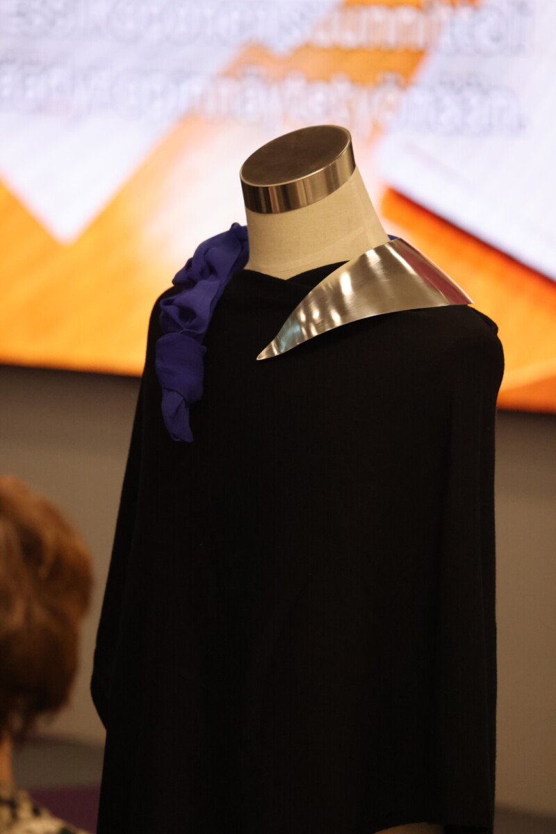 Mallinukella on musta vaate, jossa on metallinhohtoinen kaulus ja violetti röyhelöinen korostus toisella olkapäällä, oranssin ja valkoisen sävyistä sumeaa taustaa vasten.