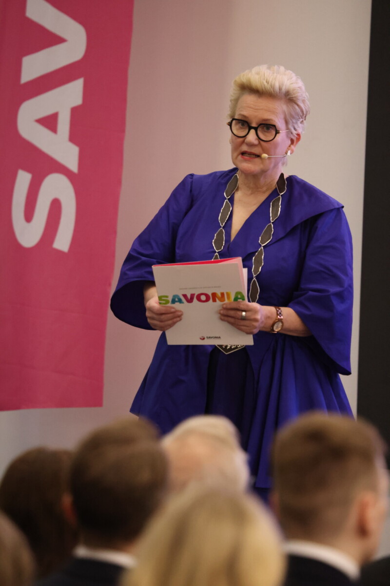 Lavalla puhuu siniseen mekkoon pukeutunut nainen, jolla on silmälasit ja suuri kaulakoru, ja hänellä on kädessään kansio, jossa lukee Savonia. Taustalla näkyy vaaleanpunainen banderolli, jossa on osittain Savonia-teksti. Etualalla on yleisön päitä.