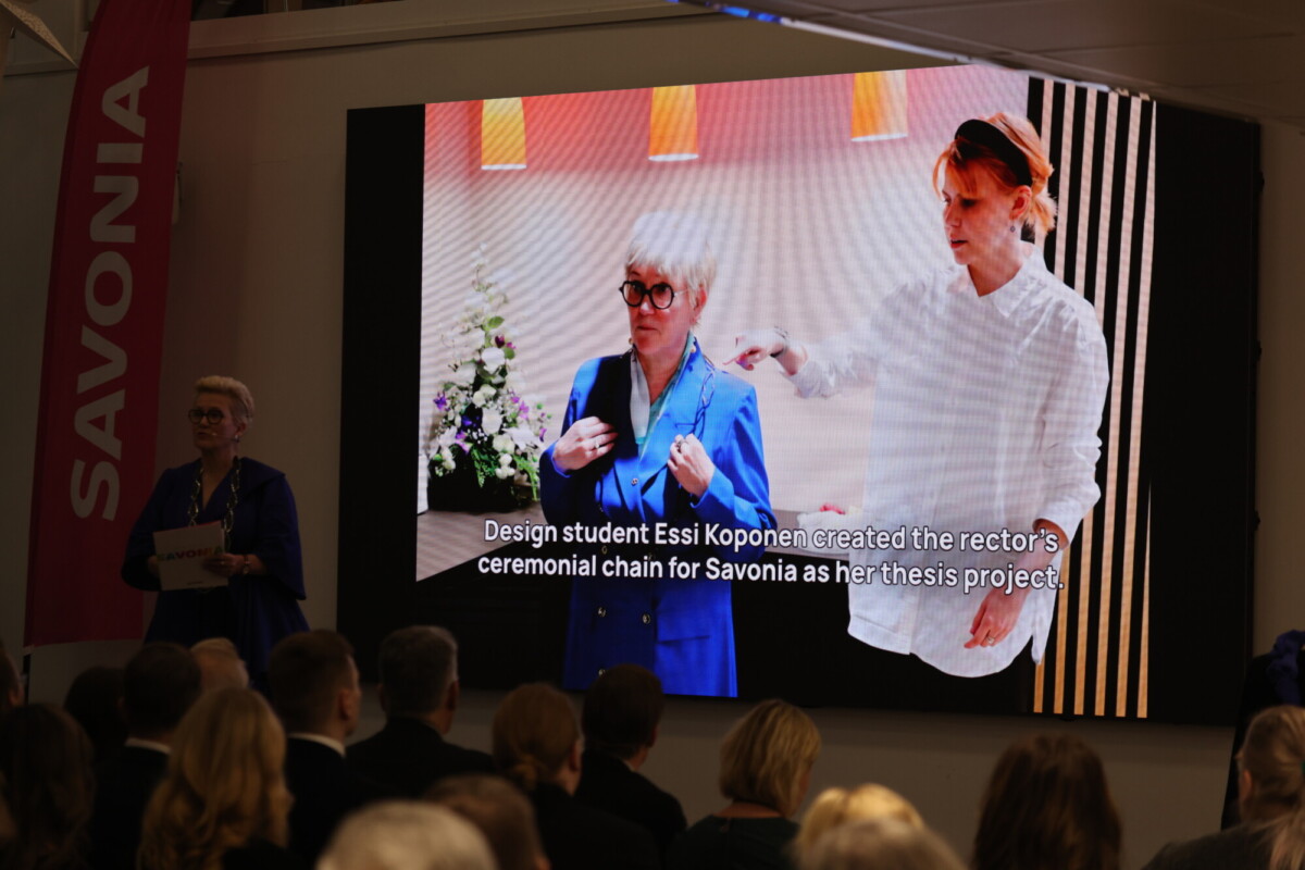 Suurella näytöllä näkyy kaksi naista, toinen sinisissä ja toinen valkoisissa vaatteissa, ja teksti erään muotoilun opiskelijan opinnäytetyöprojektista. Yleisö istuu, ja nainen seisoo lavalla vaaleanpunaisen SAVONIA-banderollin vieressä.