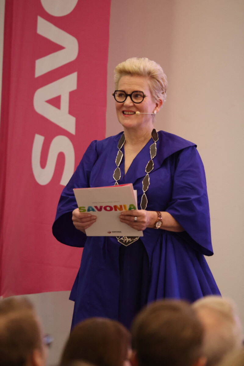 Siniseen kaapuun ja silmälaseihin pukeutunut nainen seisoo lavalla hymyillen, kädessään Savonia-kansio. Taustalla roikkuu vaaleanpunainen banderolli, jossa on valkoisin kirjaimin Savonia. Etualalla näkyy yleisön jäseniä.