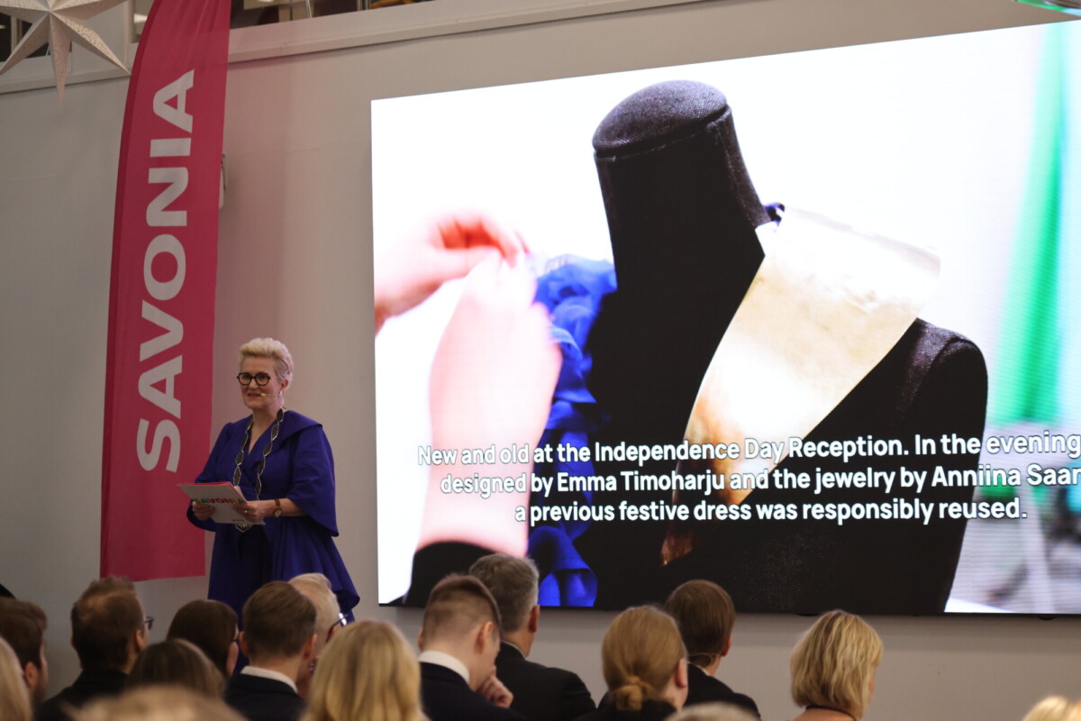 Nainen puhuu lavalla yleisön edessä. Hänen takanaan on suuri valkokangas, jossa näkyy lähikuva käsistä, jotka työskentelevät mallinuken päällä olevan mekon parissa, ja teksti, jossa kuvataan uudelleen käytettävää juhlamekkoa. Vasemmalla näkyy SAVONIA-banneri.