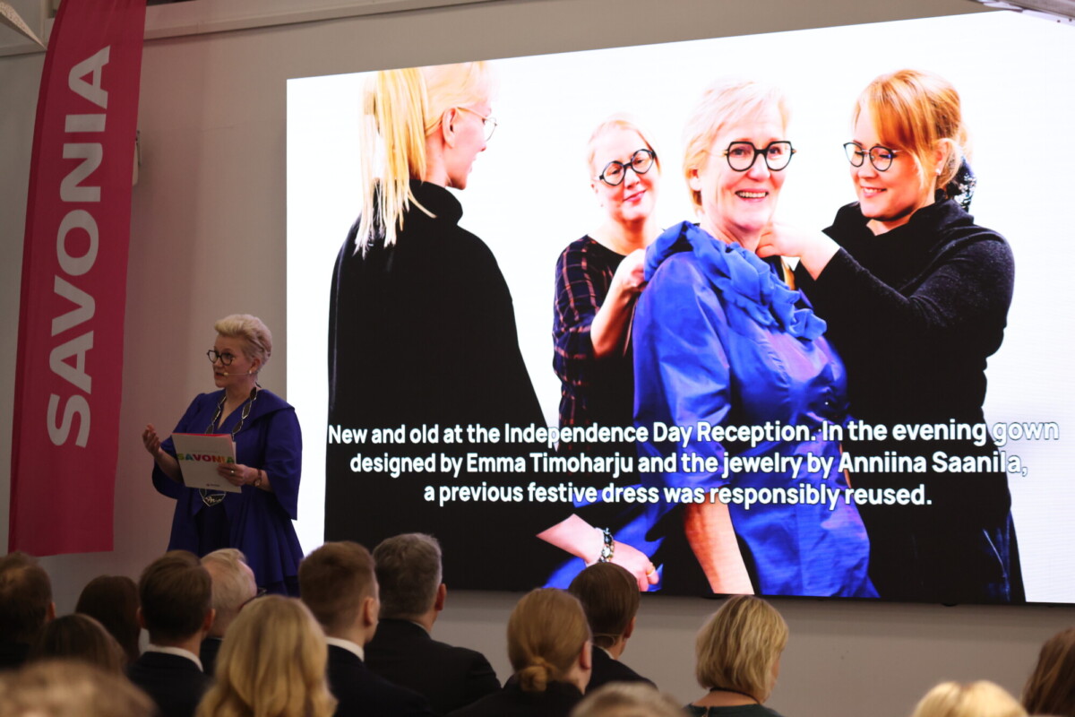Puhuja puhuu yleisölle Savonia-tapahtumassa. Hänen takanaan suurella näytöllä näkyy kuva kolmesta naisesta, jotka ihailevat sinistä pukua, ja kuvatekstissä kerrotaan puvun ja korujen kestävästä, uudelleenkäytetystä suunnittelusta.