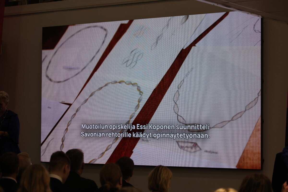 Suurella valkokankaalla näytetään paperille piirrettyjä korujen luonnoksia suomenkielisin tekstityksin. Yleisö istuu valkokankaan edessä ja seuraa esitystä hämärässä huoneessa.