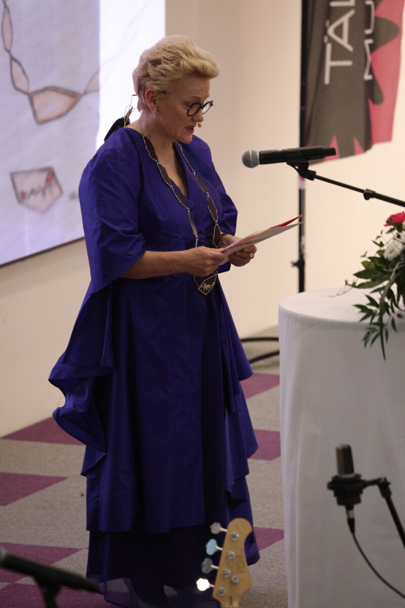 Siniseen mekkoon pukeutunut nainen seisoo korokkeella ja lukee kortista mikrofoniin. Hänellä on silmälasit ja kaulakoru, ja hänen lähellään näkyy kukka-asetelma ja mustavalkoinen banderolli.