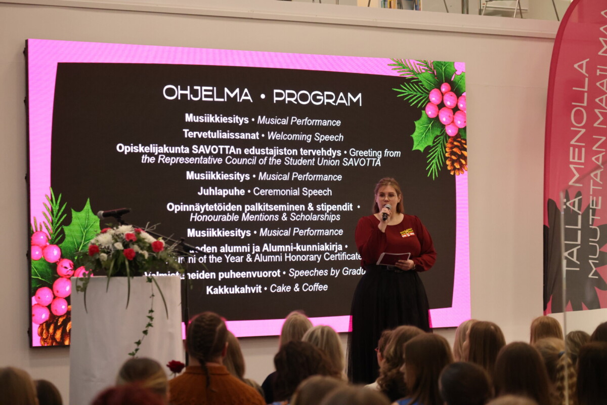 Nainen seisoo lavalla mikrofoni kädessään ja näytön edessä on kaksikielinen tapahtumaohjelma. Etualalla istuu yleisö, ja ruudulla näkyy juhlallisia koristeita.