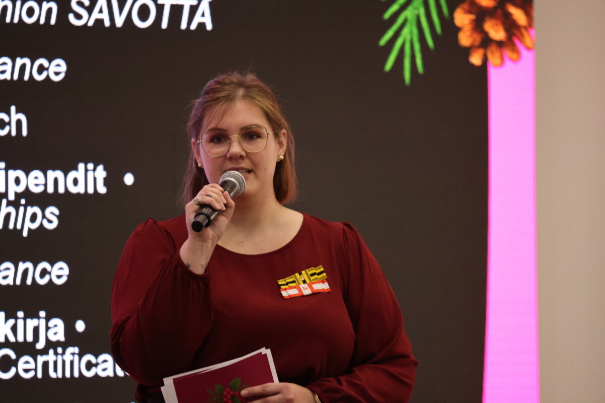 Vaaleanruskeat hiukset ja silmälasit päässä oleva nainen puhuu mikrofoniin, kädessään papereita. Hänellä on ruskeankeltainen toppi, jossa on pieniä lipputappeja, ja hän seisoo valkokankaalla, jossa on valkoista tekstiä ja kukkagrafiikkaa.