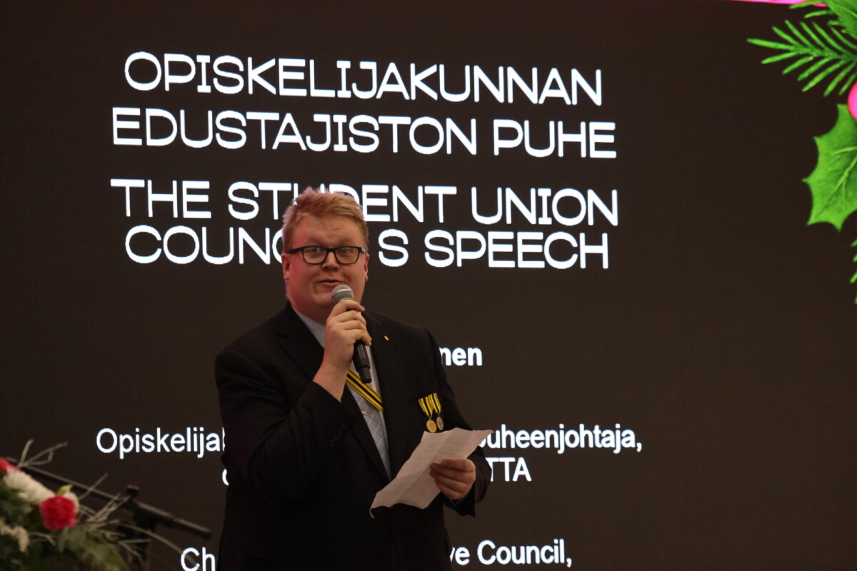 Pukuun ja solmioon pukeutunut henkilö pitää kädessään mikrofonia ja paperia ja puhuu lavalla näytön edessä, jossa näkyy tekstiä suomeksi ja englanniksi. Näytön reunalla näkyy juhlallisia koristeita.