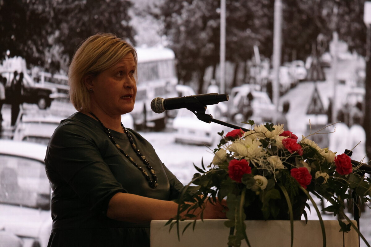 Lyhyet vaaleat hiukset omaava nainen puhuu mikrofoniin valkoisilla ja punaisilla kukilla koristellulla korokkeella, taustalla mustavalkoinen kaupunkikuvio.