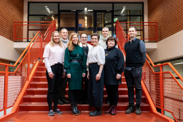 Kahdeksan hengen ryhmä seisoo yhdessä punaisilla portailla sisätiloissa ja hymyilee kameralle. Taustalla on tiiliseinät, kaiteet ja oviaukko portaiden yläpäässä.