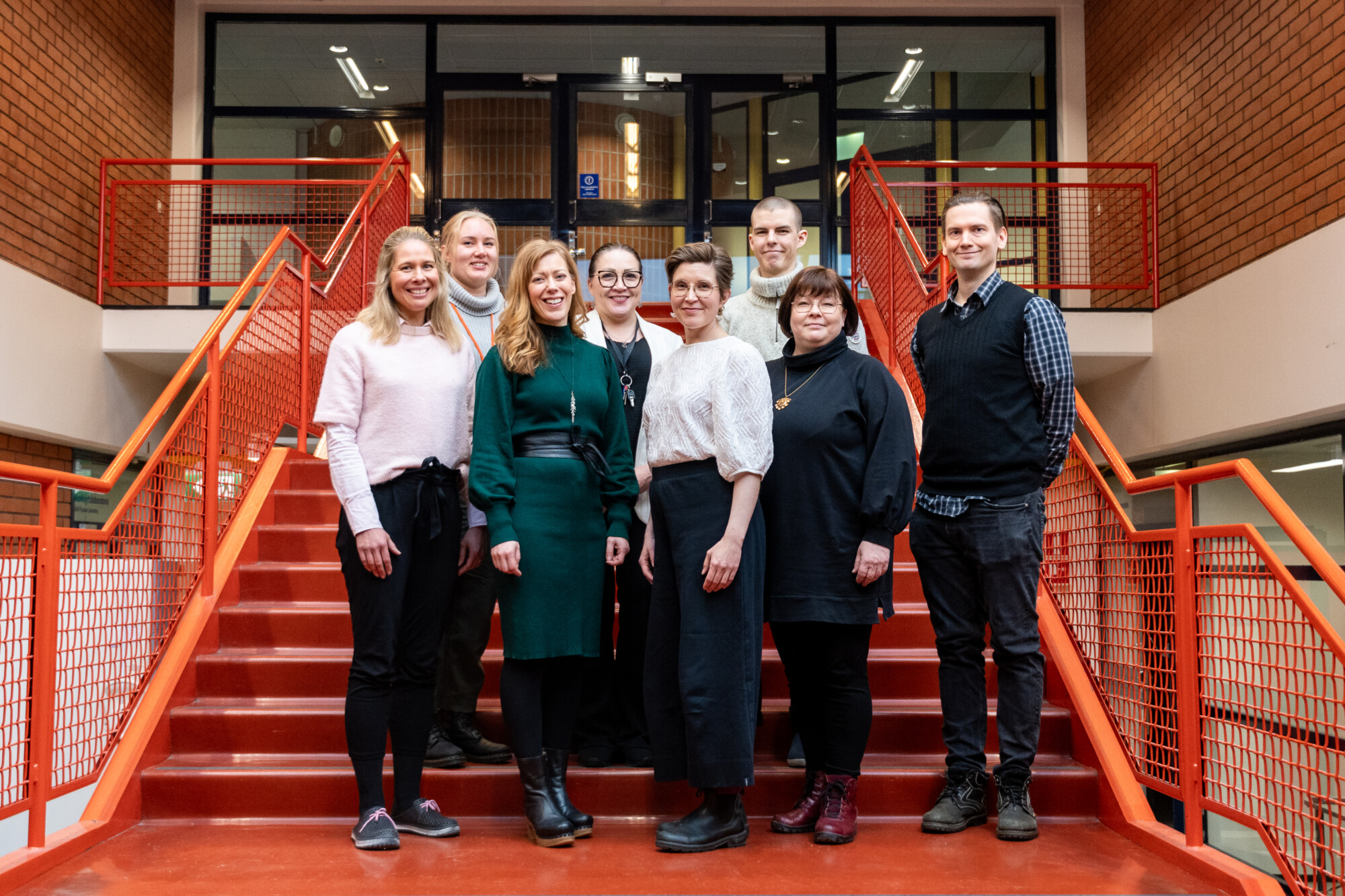 Kahdeksan hengen ryhmä seisoo yhdessä punaisilla portailla sisätiloissa ja hymyilee kameralle. Taustalla on tiiliseinät, kaiteet ja oviaukko portaiden yläpäässä.