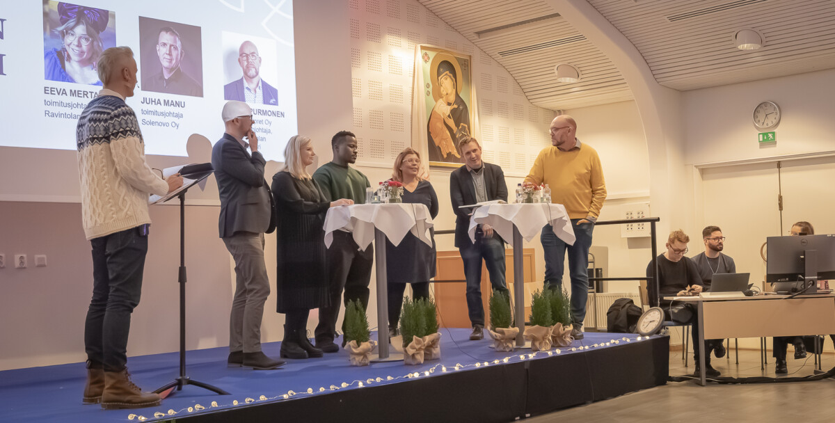 Seitsemän ihmistä seisoo lavalla ja käy paneelikeskustelua moderaattorin kanssa, ja muut istuvat läheisissä pöydissä. Taustalla näkyy suuri muotokuva ja esityksen diat. Lavalla on pieniä koristeltuja pöytiä ja viherkasveja.