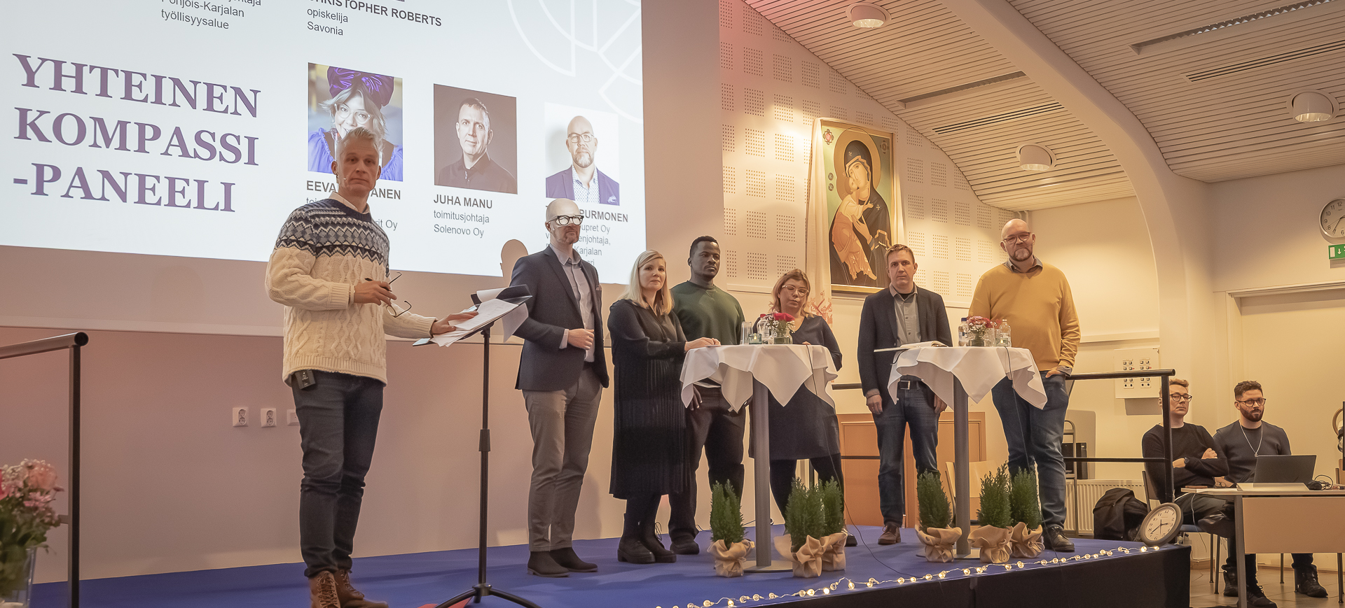 Seitsemän ihmistä seisoo lavalla paneelitilaisuuden aikana, ja heidän takanaan on valkokangas, jossa näkyy heidän nimensä. Heidän edessään on kolme valkoista pyöreää pöytää, joissa on kukkia ja kynttilöitä, ja oikealla näkyy yleisö.