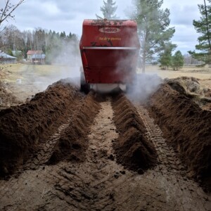 Punainen maatalouskone liikkuu eteenpäin ja luo kaksi samansuuntaista riviä muokattua maata, jossa on näkyvät jäljet ja höyryn nousu, ulkona puiden ja pellon läheisyydessä pilvisen taivaan alla.