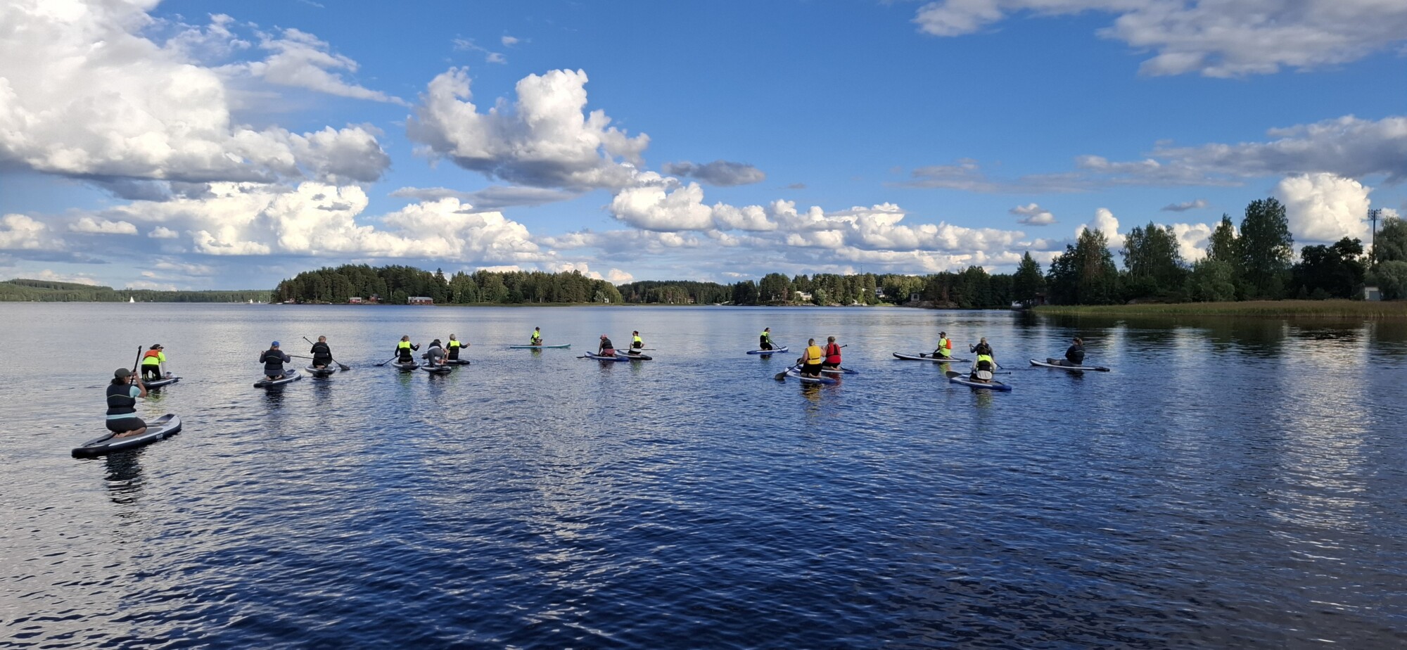 Joukko ihmisiä stand up -melontalaudoilla on kokoontunut rauhalliselle järvelle sinisen taivaan alle, jossa on hajapilviä, puiden ja kaukaisen rantaviivan ympäröimänä.
