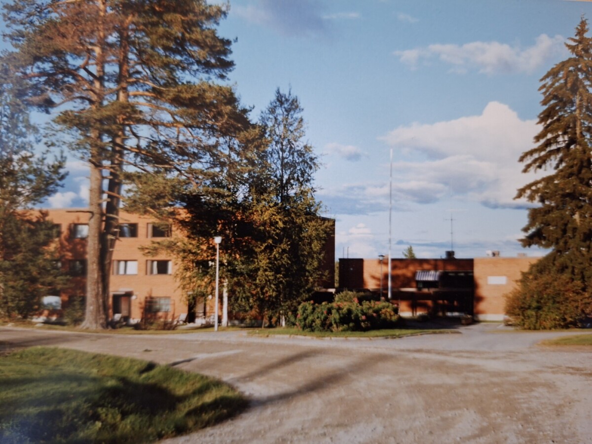 Tiilirakennus, jonka korkeat puut ovat osittain peittäneet, ja sisäänkäynnille johtaa hiekkatie sinisen taivaan alla, jossa on hajapilviä.