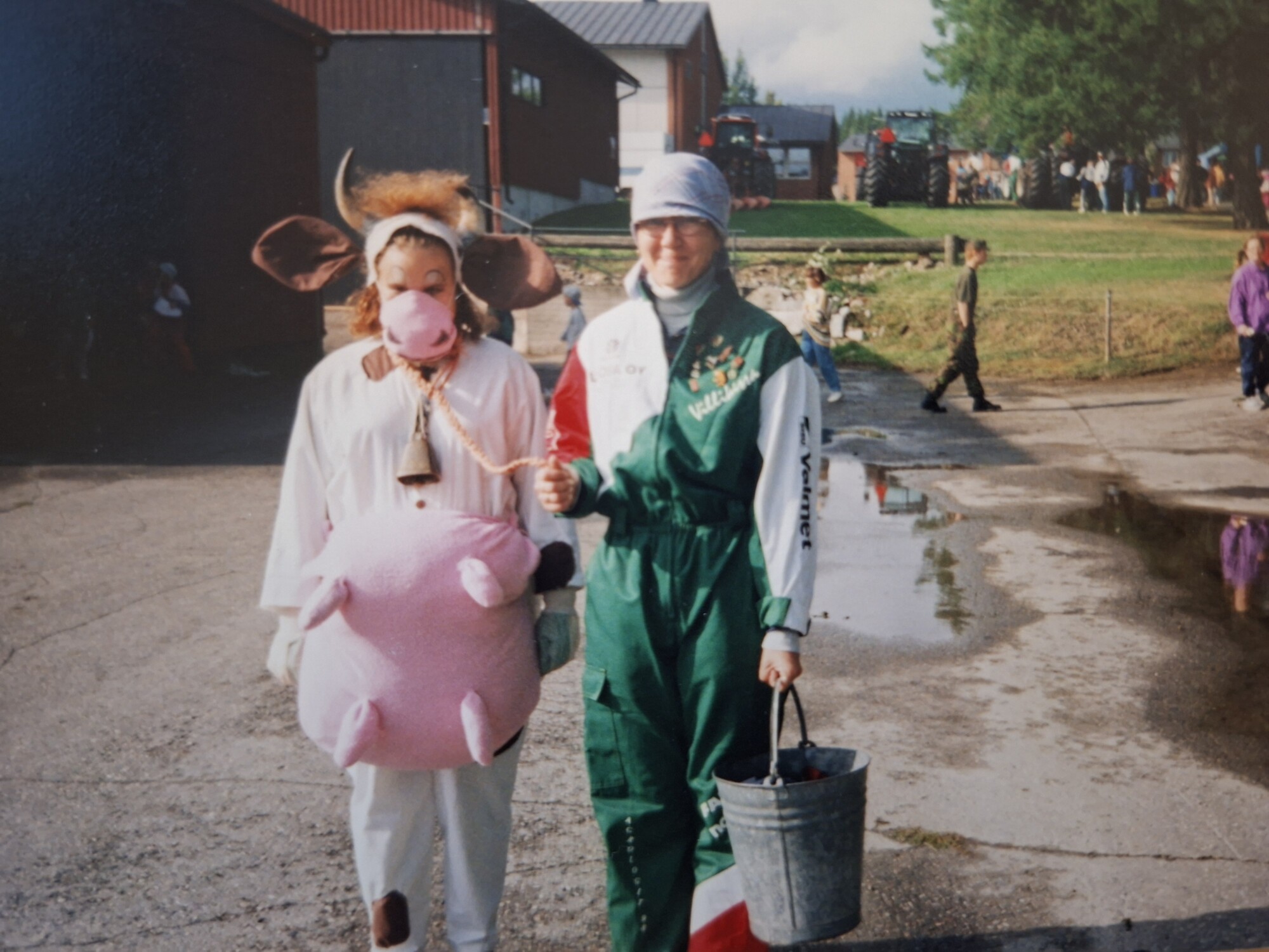 Kaksi ihmistä ulkoilmatapahtumassa; toinen on pukeutunut lehmäpukuun, jossa on vaaleanpunainen utare ja naamio, kun taas toisella on vihreä haalari ja päähuivi, ja hänellä on ämpäri kädessään ja hän johtaa lehmää köyden avulla. Taustalla näkyy muita ihmisiä.