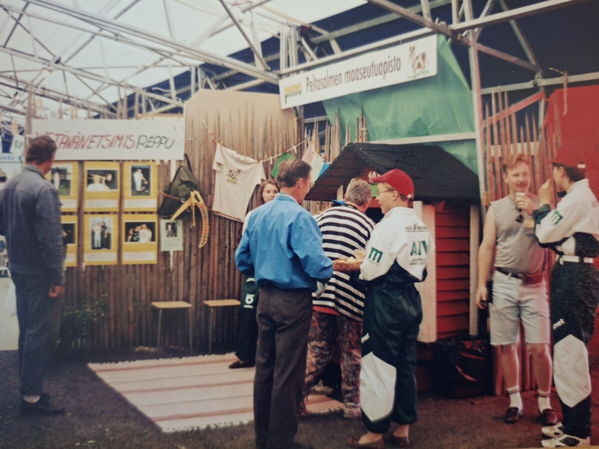 Ryhmä ihmisiä vuorovaikutuksessa ulkoilmanäyttelykojulla, jossa on esillä banderolleja ja valokuvia. Joillakin on värikkäät takit, ja yksi henkilö kättelee muiden tarkkaillessa. Kojulla on pieni puurakenne ja roikkuvia paitoja.