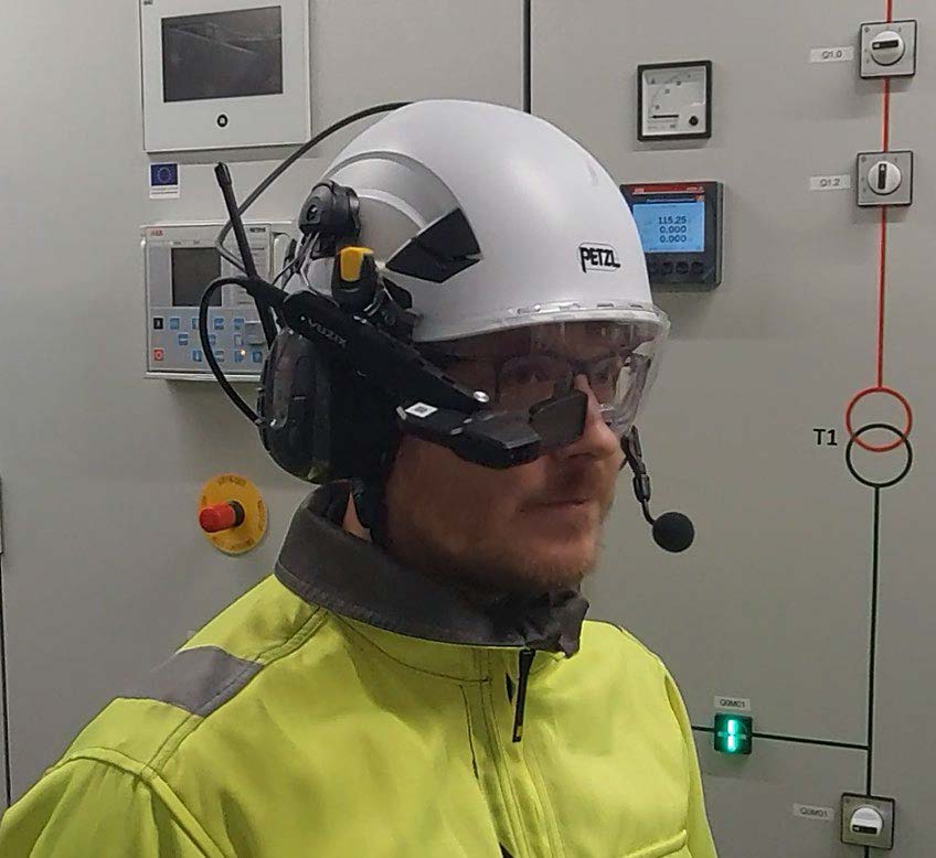 Mies, jolla on valkoinen suojakypärä, kirkkaat suojalasit ja kuulokkeet, joissa on mikrofoni ja kiinnitetyt älylasit, seisoo sähköisten ohjauspaneelien edessä. Hän on pukeutunut keltaiseen näkyvyystakkiin.