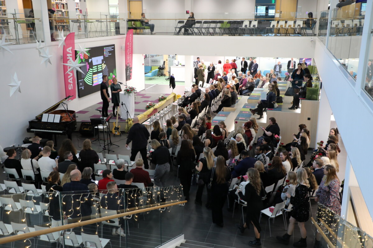 Suuri joukko ihmisiä istuu modernissa, monitasoisessa sisätilassa, jonka etuosassa on näyttämö. Tilaisuudessa on puhujia, piano, koristeita ja valonjohtoja portaita pitkin. Jotkut ihmiset sekoittuvat ja seisovat.