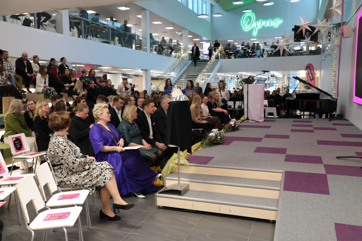 Suuri joukko ihmisiä istuu lavaa vastapäätä modernissa rakennuksessa, jossa on korkeat katot ja lasikaiteet. Yleisöön kuuluu miehiä ja naisia, joista osa on pukeutunut muodollisesti. Lavalla on kukkia ja lähellä on flyygeli.