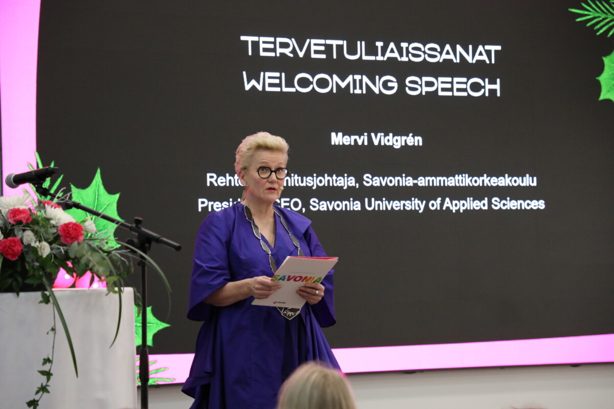 Nainen seisoo korokkeella papereita kädessään ja puhuu tilaisuudessa suomenkielisen ja englanninkielisen tervehdyspuheen näytön edessä. Korokkeen vasemmalla puolella näkyy kukka-asetelma.