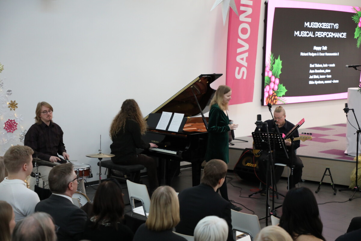 Pieni yhtye esiintyy lavalla pianistin, rumpalin, kitaristin ja laulajan kanssa yleisön edessä. Taustalla olevalla näytöllä näkyy "MUSIIKKIESITYS MUSIIKKIESITYS". Vaaleanpunaisia ja valkoisia koristeita on näkyvissä.