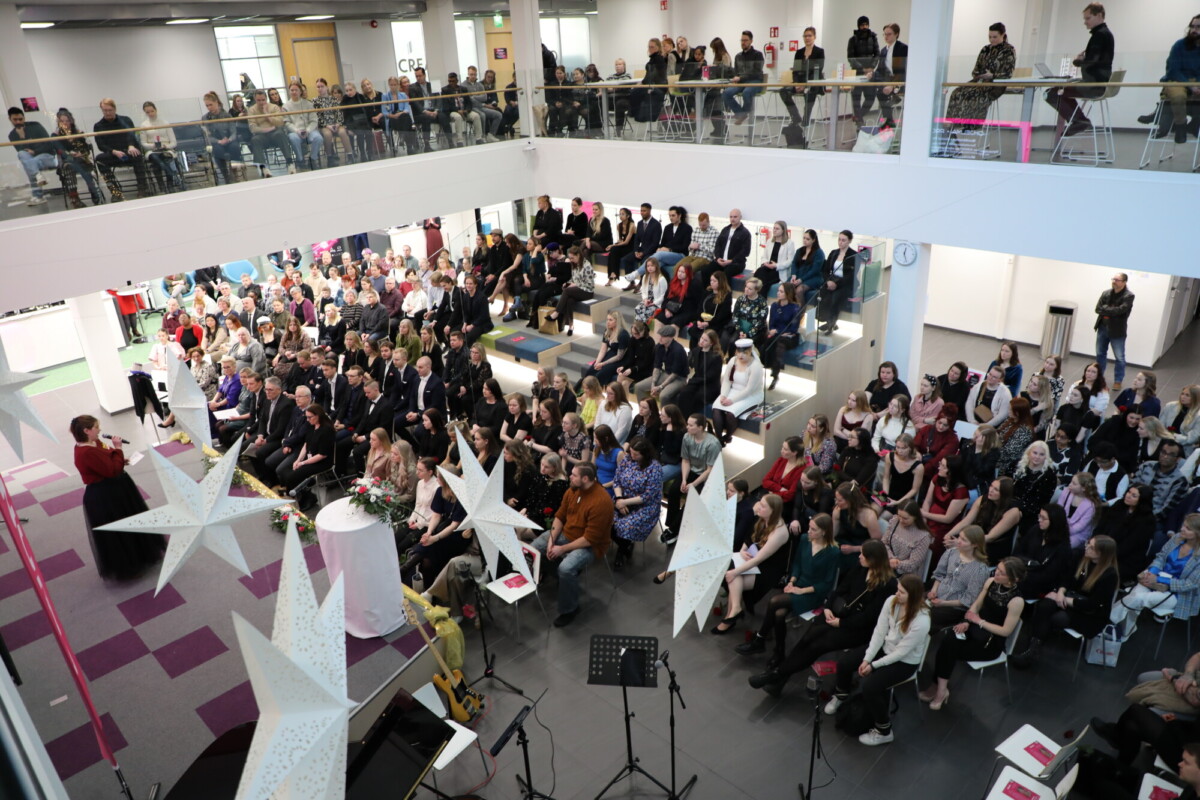 Suuri yleisö istuu modernissa, avoimessa kaksikerroksisessa rakennuksessa osallistumassa juhlatilaisuuteen. Koristeelliset valkoiset tähtilyhdyt roikkuvat edessä, ja puhuja puhuu yleisölle korokkeelta.