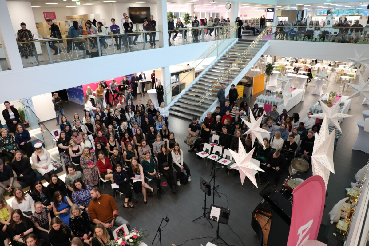 Suuri joukko ihmisiä istuu ja seisoo modernissa, avoimessa atriumissa, jossa on korkeat katot ja portaikko. Katosta roikkuu valkoisia tähtikoristeita. Istuvan yleisön läheisyyteen on pystytetty joitakin soittimia.