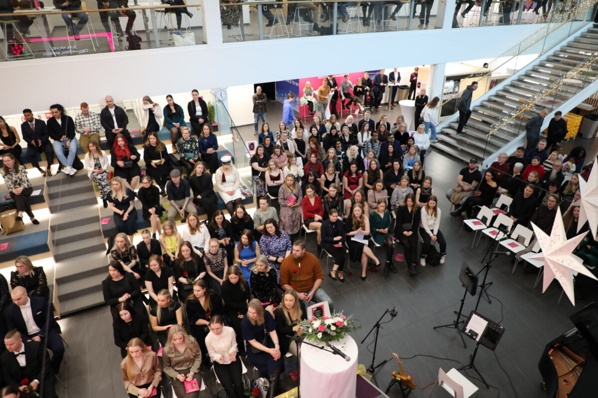 Suuri joukko ihmisiä istuu ja seisoo modernissa, kirkkaasti valaistussa atriumissa, jossa on kukkien koristama koroke. Jotkut istuvat portailla, toiset tuolirivissä. Oikealla näkyy portaikko ja koristeita.