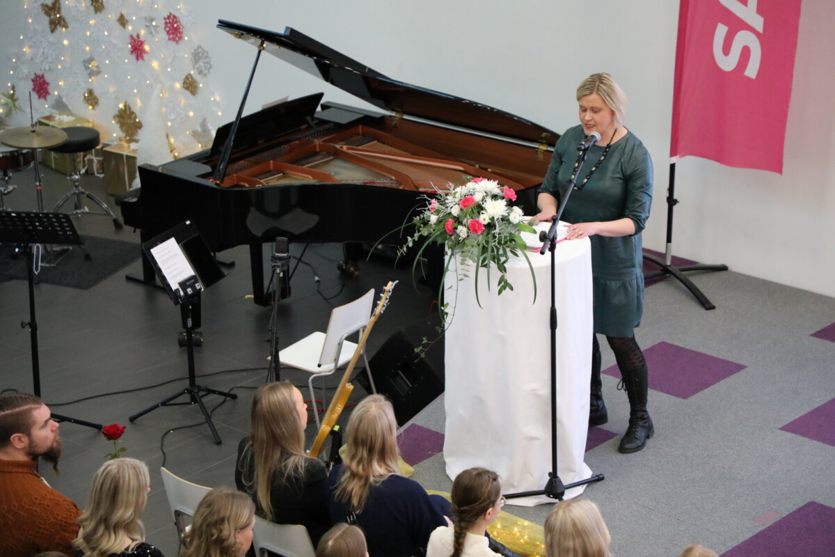 Nainen seisoo kukkien koristelemalla korokkeella ja puhuu yleisölle. Lähistöllä näkyy piano, rumpukalusto ja soittimia. Useat ihmiset istuvat ja kuuntelevat tarkkaavaisesti.