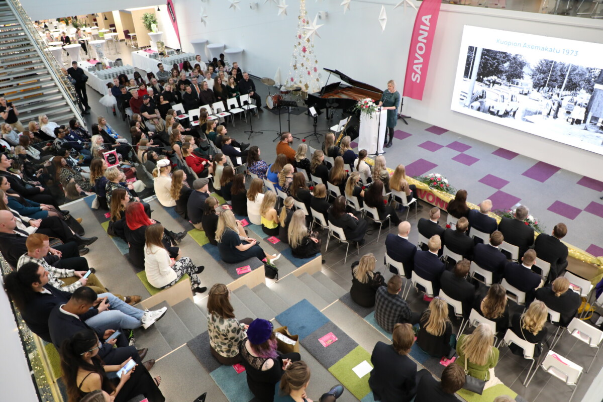 Suuri joukko ihmisiä istuu lavaa vastapäätä modernissa, valoisassa salissa. Puhuja seisoo korokkeella pianon vieressä ja esittää mustavalkoista valokuvaa valkokankaalla. SAVONIA näkyy banderollissa.