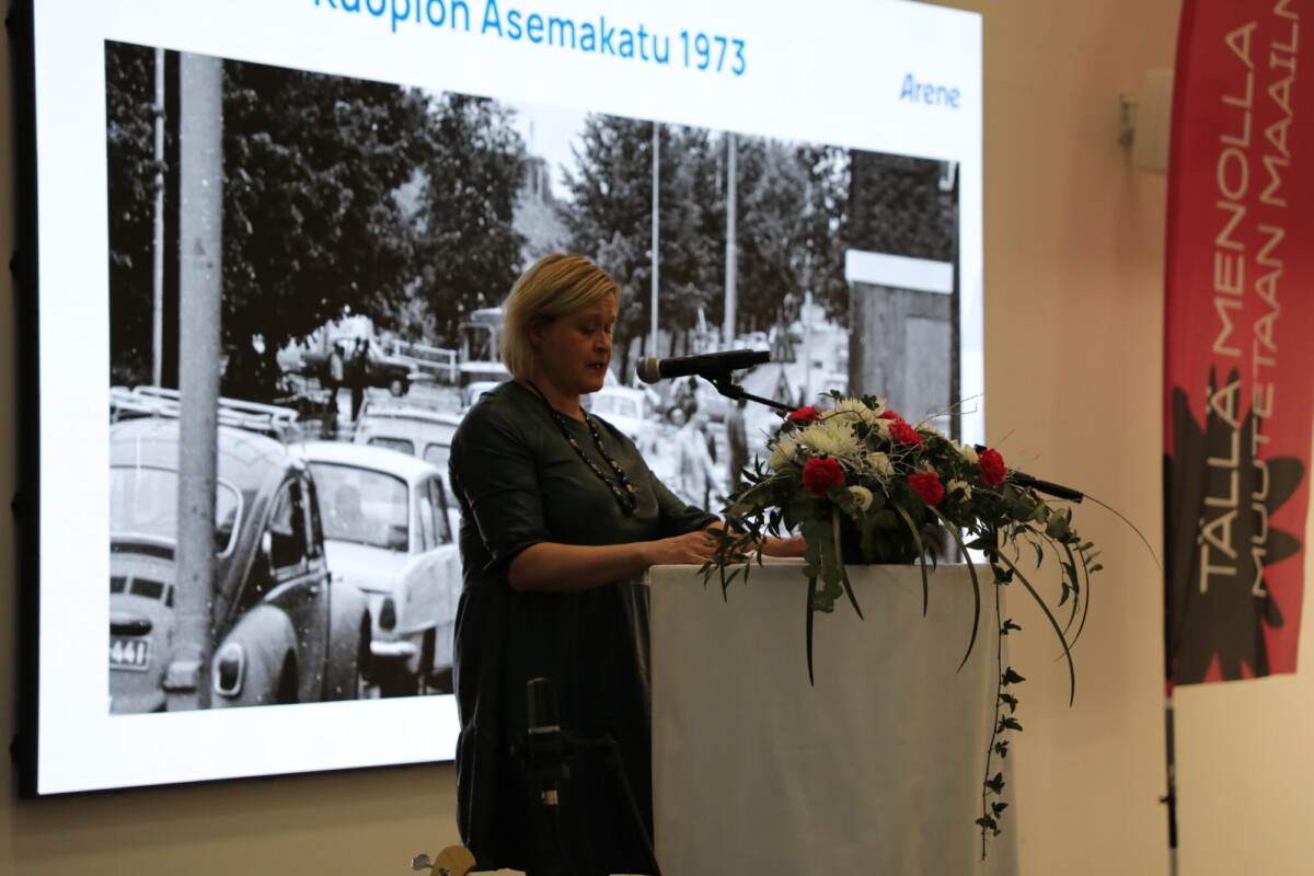 Nainen seisoo korokkeella kukkien kanssa ja puhuu mikrofoniin. Hänen takanaan on mustavalkoinen valokuva Raution Asemakatu 1973, jossa näkyy vanhoja autoja. Oikealla näkyy pystysuora vaaleanpunainen banderolli.
