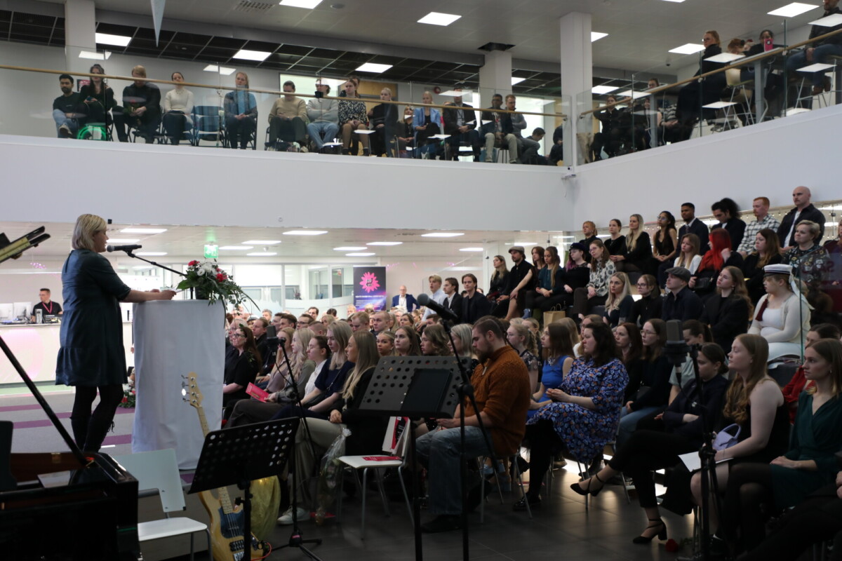 Nainen puhuu korokkeella suuren yleisön edessä, joka istuu ja seisoo modernissa, kaksitasoisessa sisätilassa. Ihmiset kuuntelevat tarkkaavaisesti, ja etualalla näkyy soittimia.