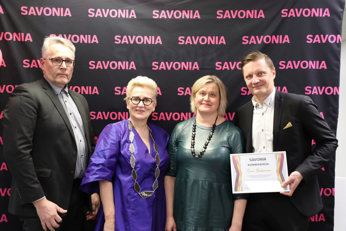 Neljä aikuista seisoo mustan taustan edessä, jossa on vaaleanpunainen SAVONIA-teksti. Yksi mies oikealla pitää kädessään todistusta, ja kaikki ovat pukeutuneet viralliseen tai puoliviralliseen asuun, poseeraavat ja hymyilevät kuvaa varten.